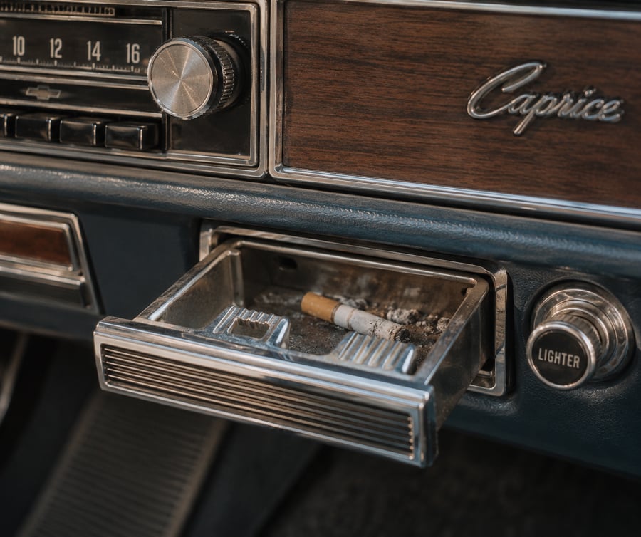 Close-up of a vintage car dashboard featuring a radio, wooden panel, lighter, and an open ashtray with a used cigarette inside.