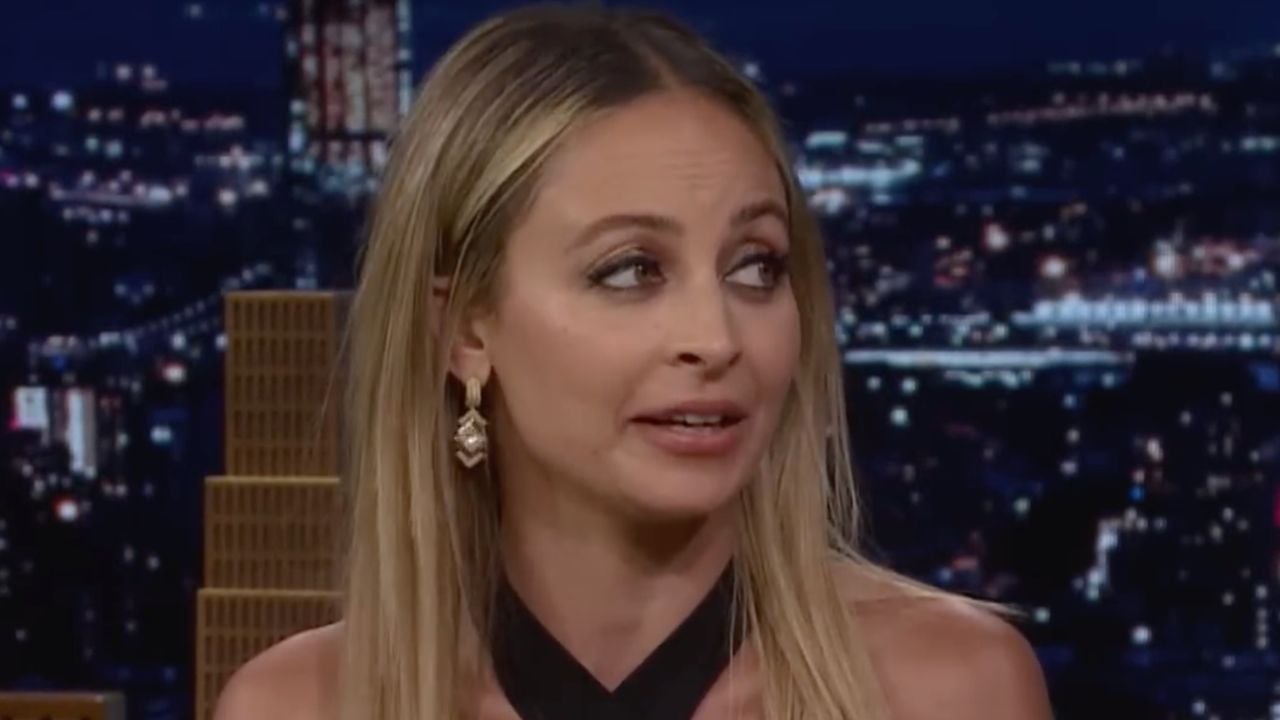 A woman with long blonde hair and dangling earrings looks to her left while speaking on a nighttime talk show, with a blurred cityscape in the background.