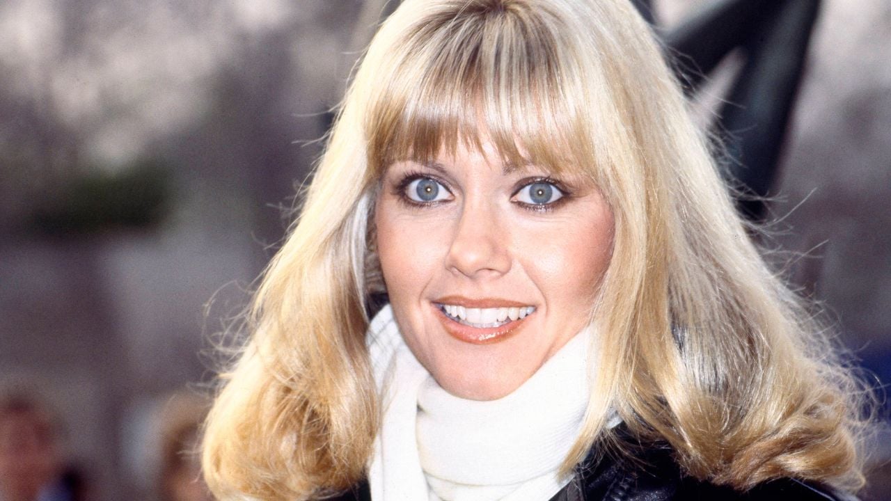 A woman with blonde hair and blue eyes smiles outdoors. She is wearing a white scarf and a black jacket.