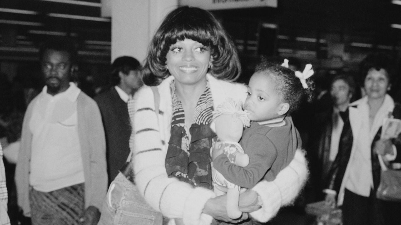 A smiling woman in a striped sweater holds a young child with bows in her hair while standing in a busy indoor public space with people in the background.
