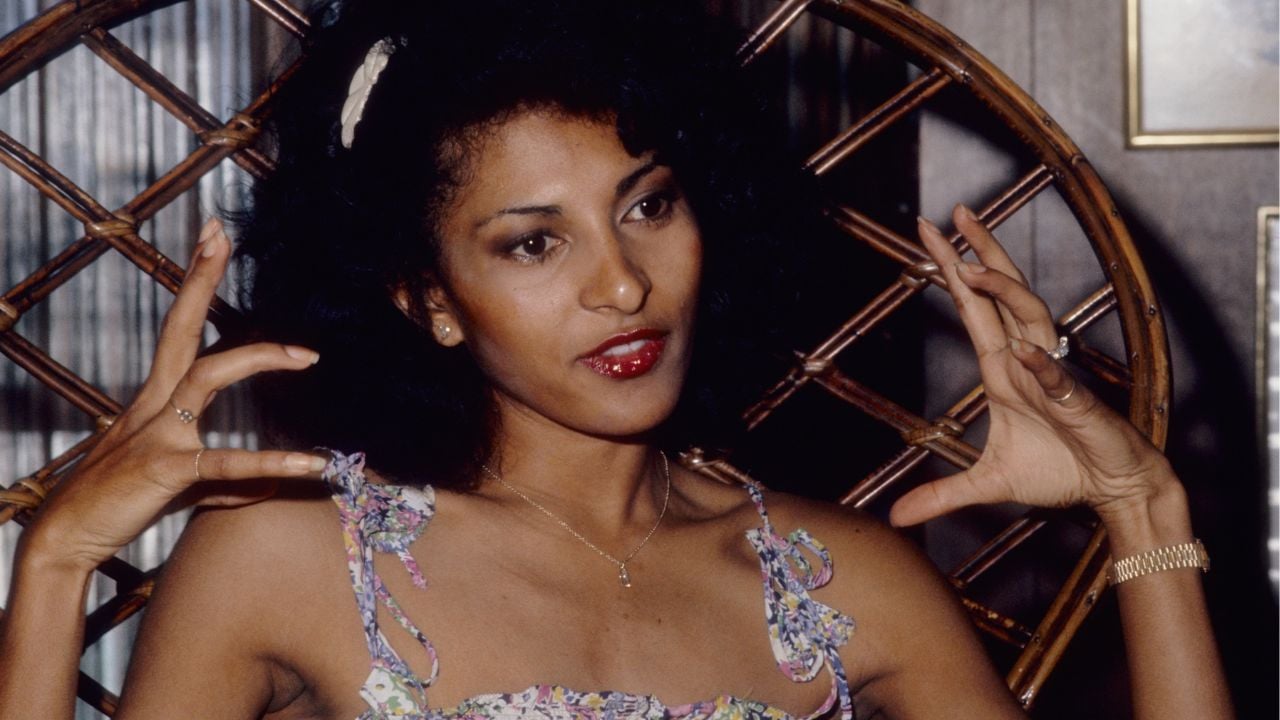 Woman with dark hair sits in a wicker chair, wearing a floral dress. She gestures with both hands near her face, looking slightly to the side, with an expressive facial expression.