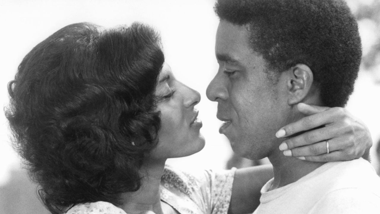 A woman with short curly hair gently holds a man’s neck with both hands as they stand close, gazing into each other’s eyes. The image is in black and white.