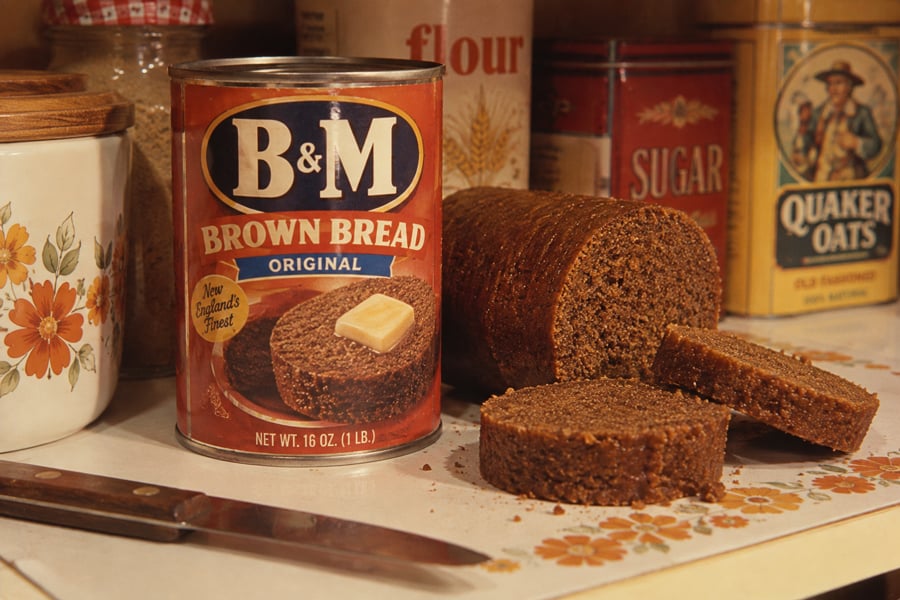 A can of B&M Brown Bread, with sliced bread next to it on a cutting board. A knife and jars labeled “flour” and “sugar” are in the background, along with a box of Quaker Oats.