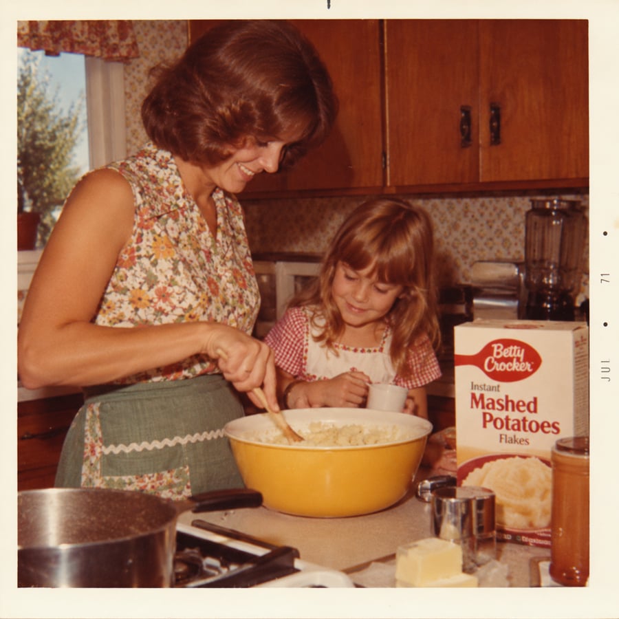 A woman and a young girl mix mashed potatoes in a yellow bowl in a kitchen. A box of Betty Crocker Instant Mashed Potatoes is on the counter. The woman wears an apron and the girl holds a measuring cup.