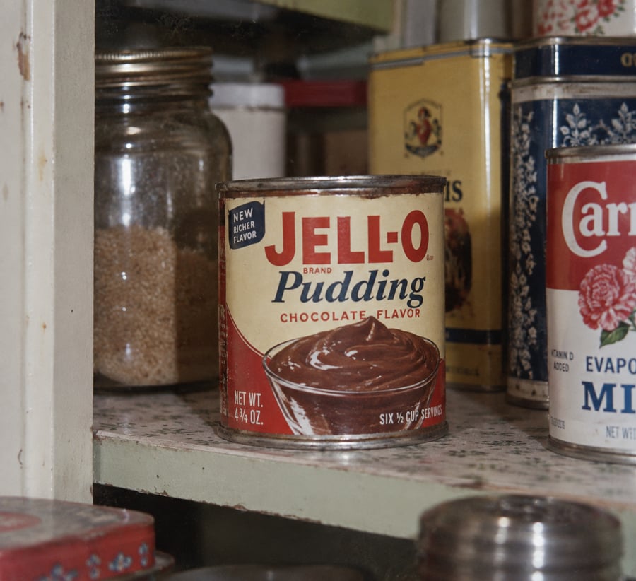 A can of Jell-O chocolate pudding sits on a pantry shelf surrounded by jars and cans, including one labeled “Carnation Evaporated Milk.”