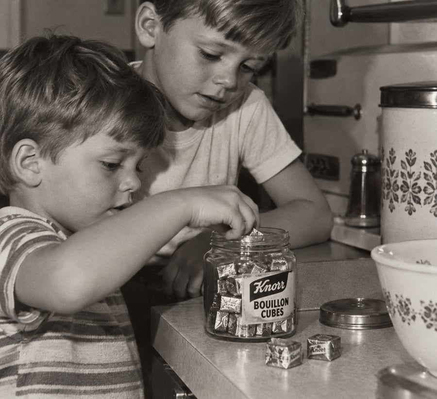 Two young boys stand in a kitchen. One boy reaches into a glass jar labeled “Knorr Bouillon Cubes” and picks up a wrapped cube, while the other watches. Several bouillon cubes are on the counter nearby.