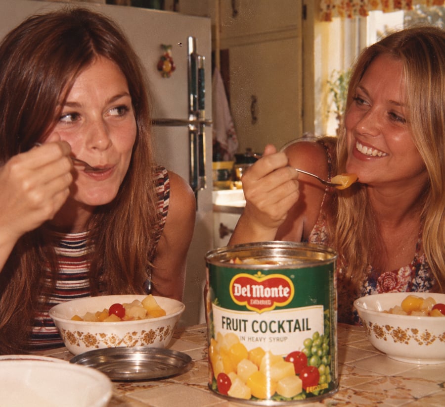 Two women smile and eat fruit cocktail from bowls at a kitchen table. An open Del Monte fruit cocktail can sits between them. The kitchen looks homey and vintage.