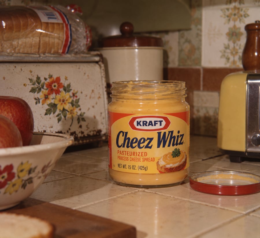 An open jar of Kraft Cheez Whiz sits on a kitchen counter near a yellow toaster, bread, apples, and vintage kitchen decor. The jar’s lid is off and resting beside it.
