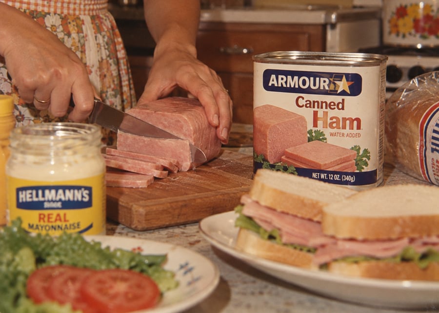 A person slices canned ham on a wooden board next to a jar of mayonnaise. Sandwiches with ham and lettuce are on a plate, with tomatoes and lettuce nearby.
