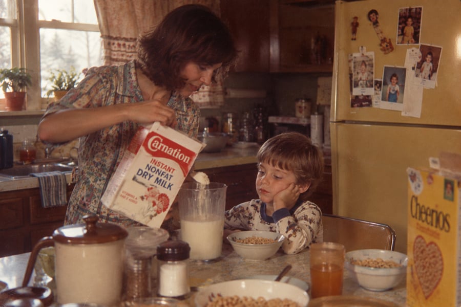 A woman pours dry milk into a pitcher at a kitchen table while a young boy watches. Bowls of cereal, a box of Cheerios, and various kitchen items are on the table. A yellow fridge is in the background.