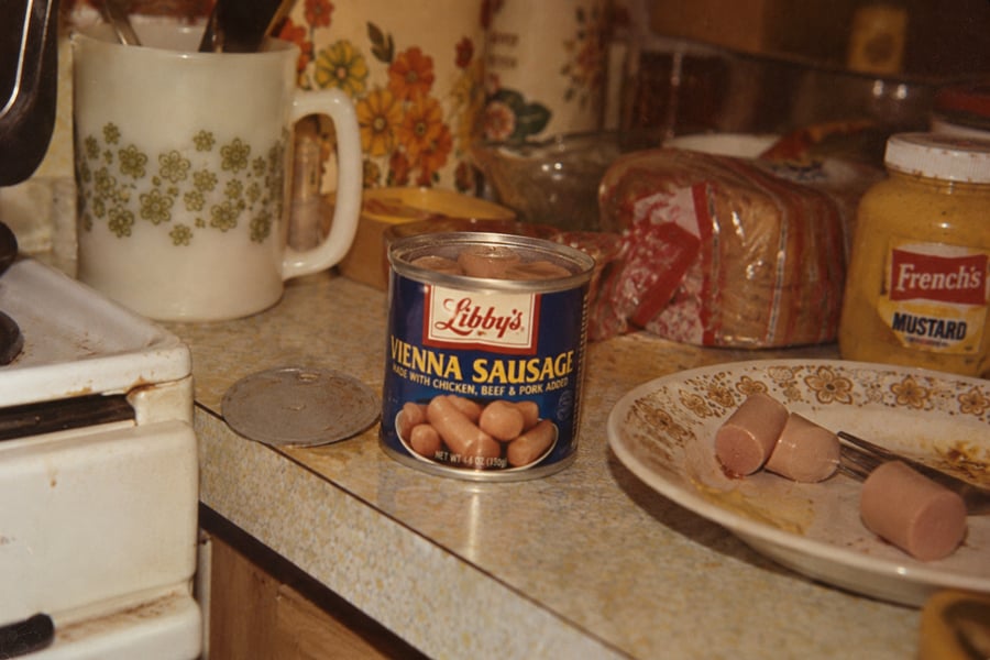 A can of Libby’s Vienna Sausage sits open on a kitchen counter, with sausages on a plate, a mug, French’s mustard, bread, and a floral plate nearby. The scene appears casual and slightly cluttered.