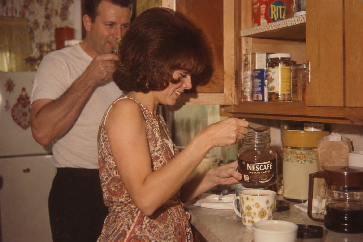 A woman makes instant coffee in a kitchen, smiling, while a man in the background sips from a cup. The counter holds coffee items and mugs, and kitchen shelves are visible.