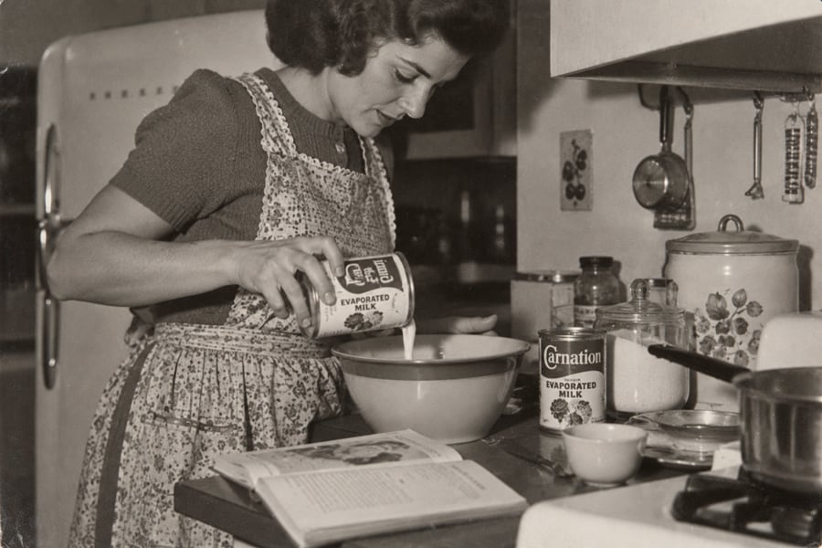 A woman in a vintage kitchen pours evaporated milk into a mixing bowl. She wears an apron and stands beside an open cookbook, with a can of Carnation milk and other ingredients on the counter.