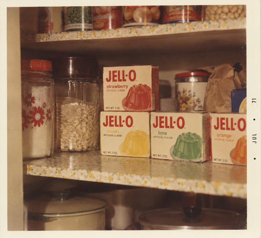 Three boxes of Jell-O (strawberry, lemon, lime, orange) sit on a pantry shelf, surrounded by jars, cans, and kitchen items. The photo has a vintage look with a July 1971 date on the border.