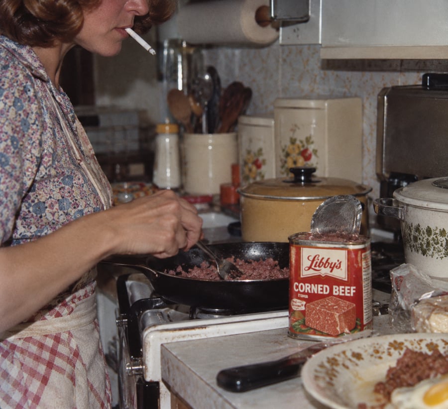 A woman cooking corned beef in a skillet on the stove, cigarette in her mouth. An open can of Libby’s Corned Beef sits nearby. Kitchen utensils and pots are in the background.