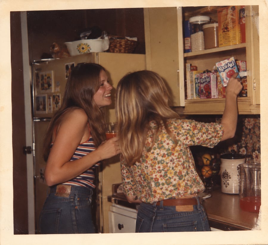 Two women in a vintage kitchen, one smiling with a drink, the other reaching for a packet of Kool-Aid from a cupboard. Both wear jeans and patterned tops; there’s a refrigerator and a pitcher on the counter.