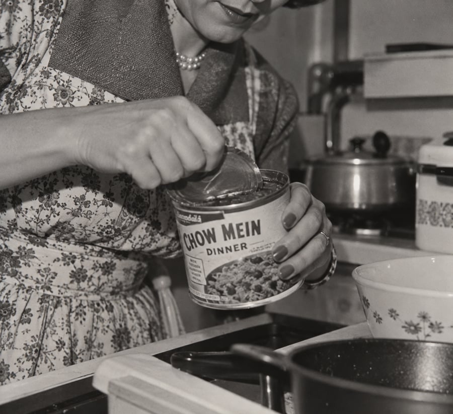 A woman in a patterned apron opens a can labeled “Chow Mein Dinner” in a vintage kitchen. Nearby are a bowl, saucepan, and kettle. Only her lower face and hands are visible.
