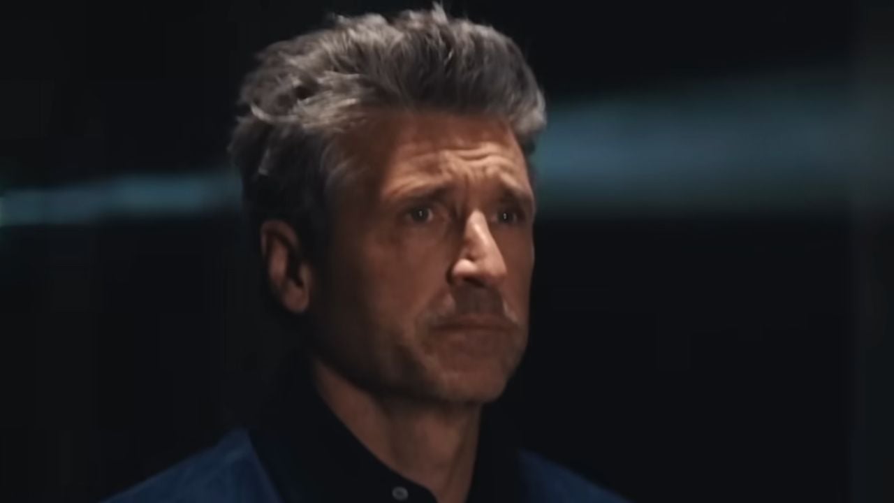A middle-aged man with gray hair and a serious expression stands in a dimly lit setting, facing forward.