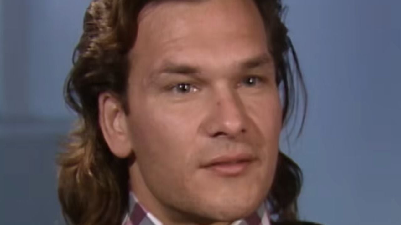 A person with long brown hair, wearing a checkered shirt and dark jacket, looks slightly off camera against a plain blue background.