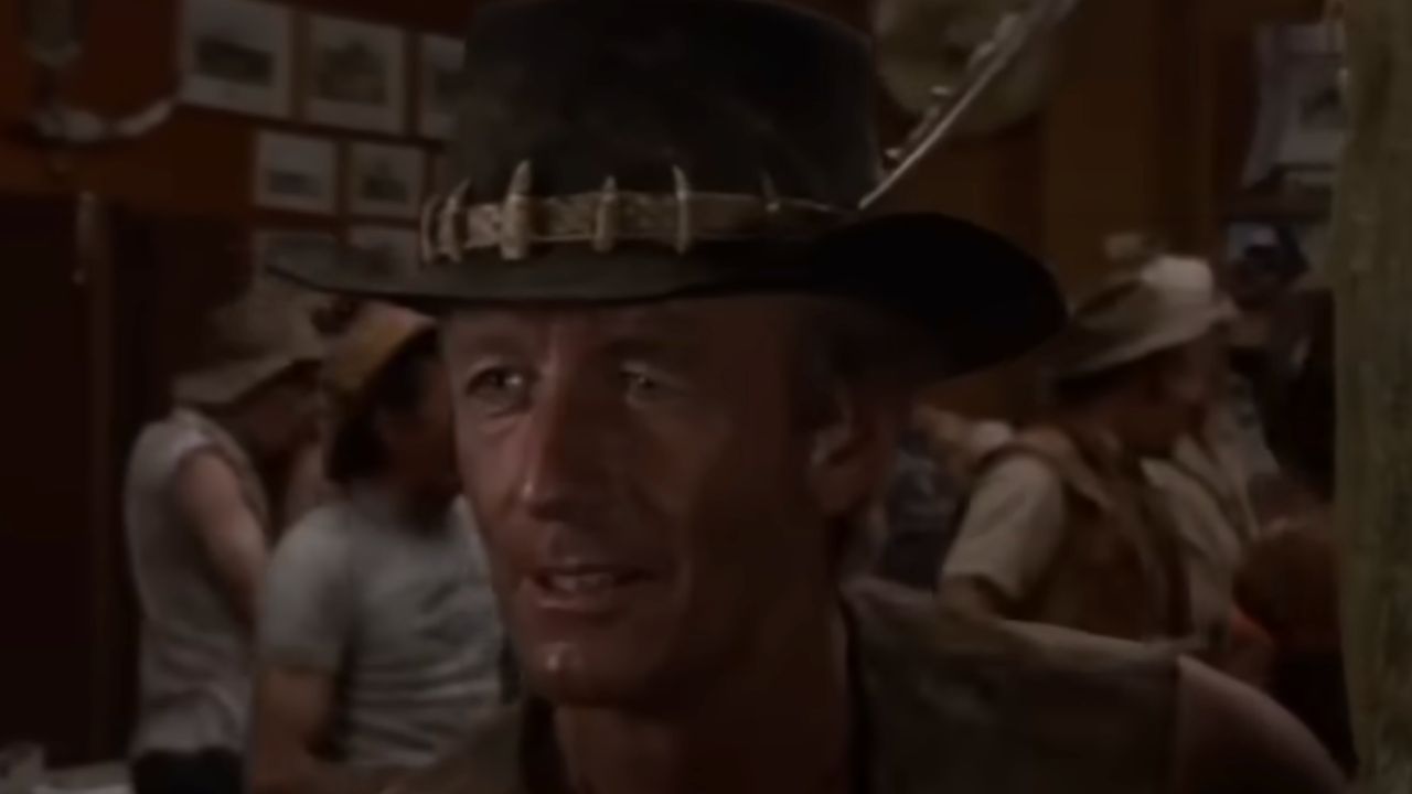 A man in a wide-brimmed hat with crocodile teeth, wearing a vest, stands in a rustic, busy bar with other people in the background.