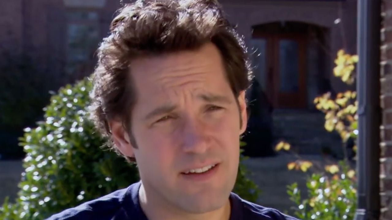 A man with short brown hair is outside, squinting slightly in the sunlight. There are green bushes and a brick building in the blurred background.