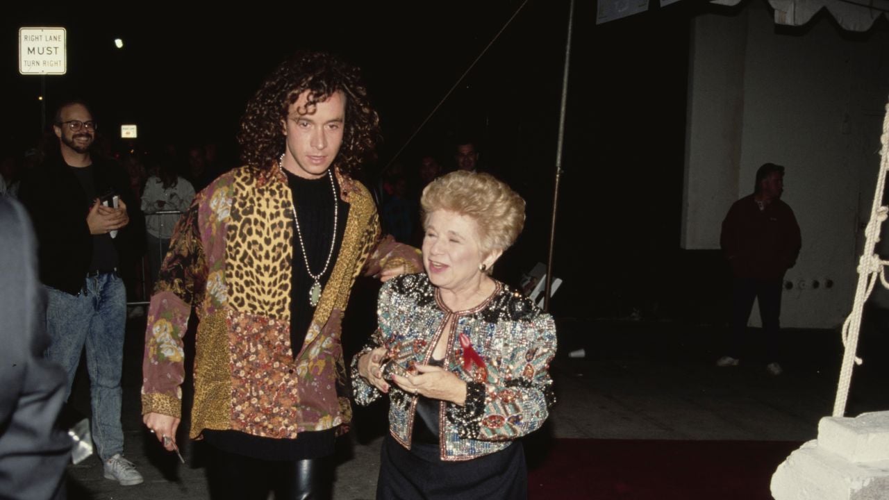 A young man in a patterned jacket walks with an older woman in a sparkling top on a red carpet at night. People and a “Right Lane Must” sign are visible in the background.