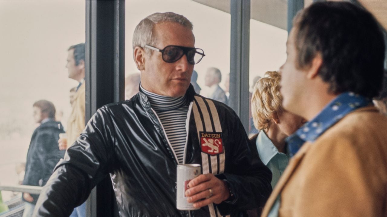 A man in sunglasses and a black racing jacket holds a drink can while talking to another man in a tan jacket indoors, with people and windows in the background.