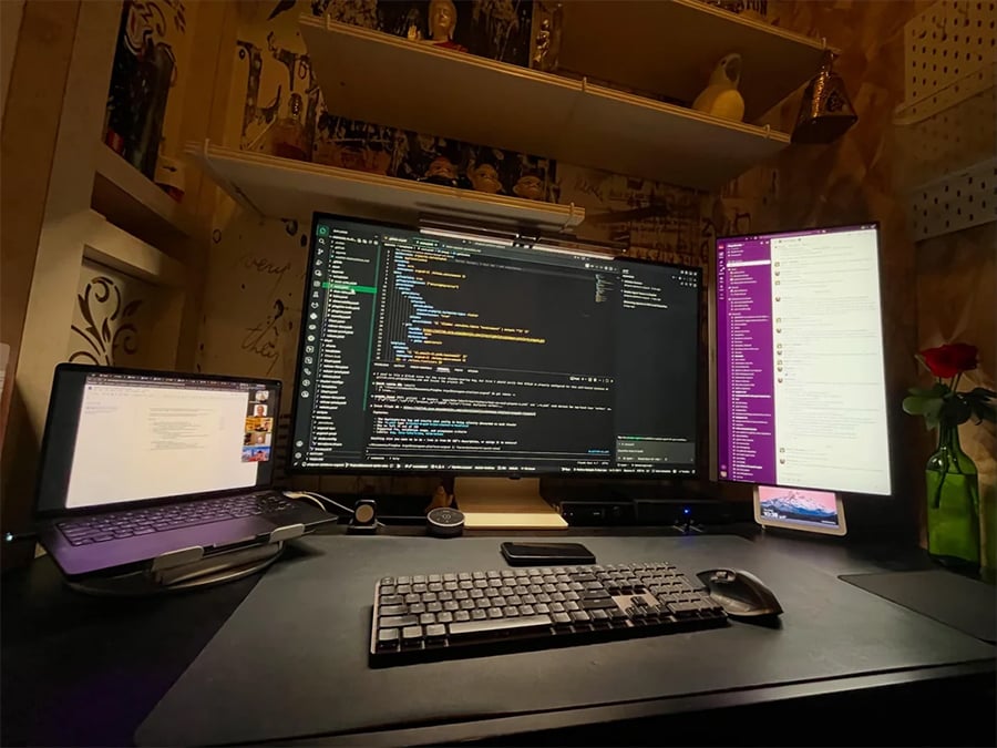 A desk setup with a laptop, a large monitor displaying code, a vertical monitor showing a chat app, a keyboard, mouse, phone, and a green vase with a red rose. Shelves with decor are in the background.