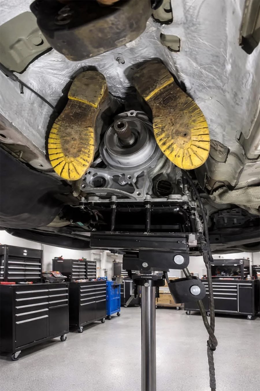 A pair of yellow work boots are humorously placed inside the undercarriage of a raised car in an auto repair shop, making it appear as if someone is trapped inside the vehicle.