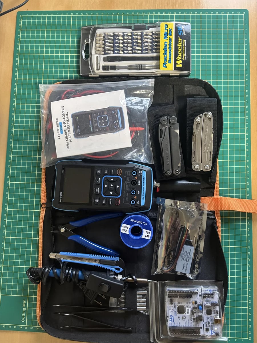 A black toolkit with an electronics tester, pliers, screwdriver set, wire, multitool, cables, pliers, and electronic components on a green cutting mat.