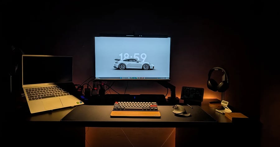 A minimalist desk setup features a desktop monitor with a car wallpaper, an open laptop, mechanical keyboard, mouse, headphones on a stand, and other accessories, all illuminated by warm ambient lighting.