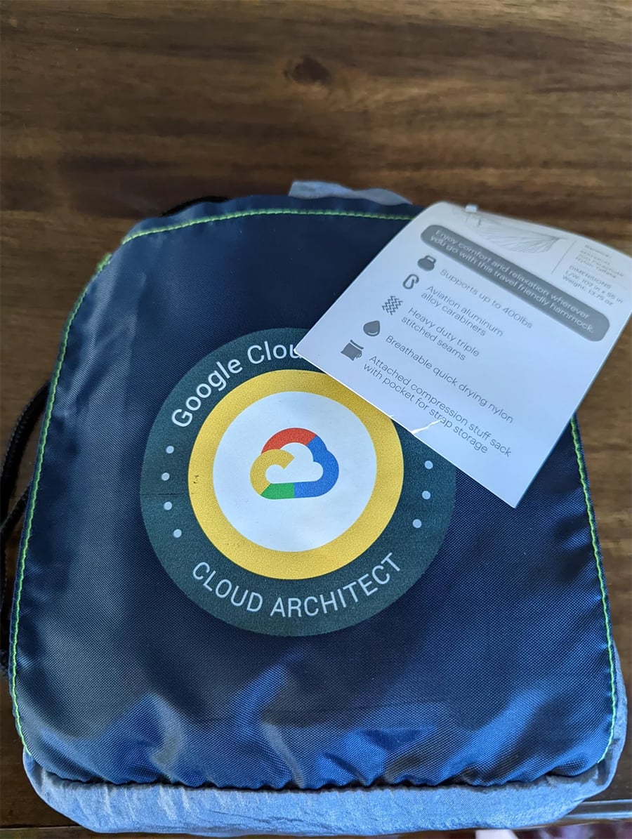 A Google Cloud Architect branded bag sits on a wooden surface, with a tag attached that describes its features, including being made of recycled nylon and having an attached storage sack.