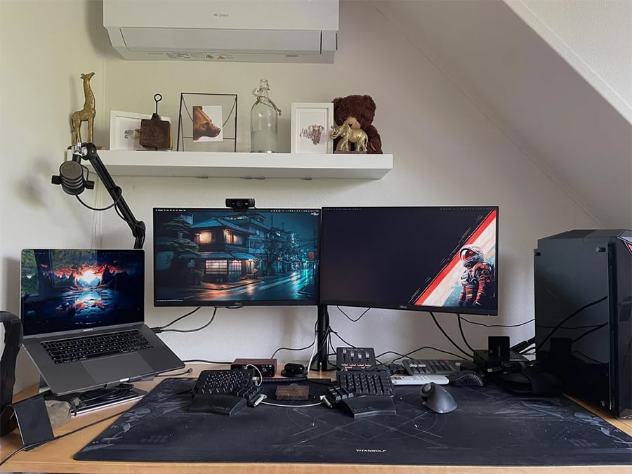 A modern desk setup with two monitors, a laptop, keyboard, mouse, gaming controllers, and desk accessories. Above, a white shelf holds art, bottles, animal figures, and a teddy bear. An air conditioner is on the wall.