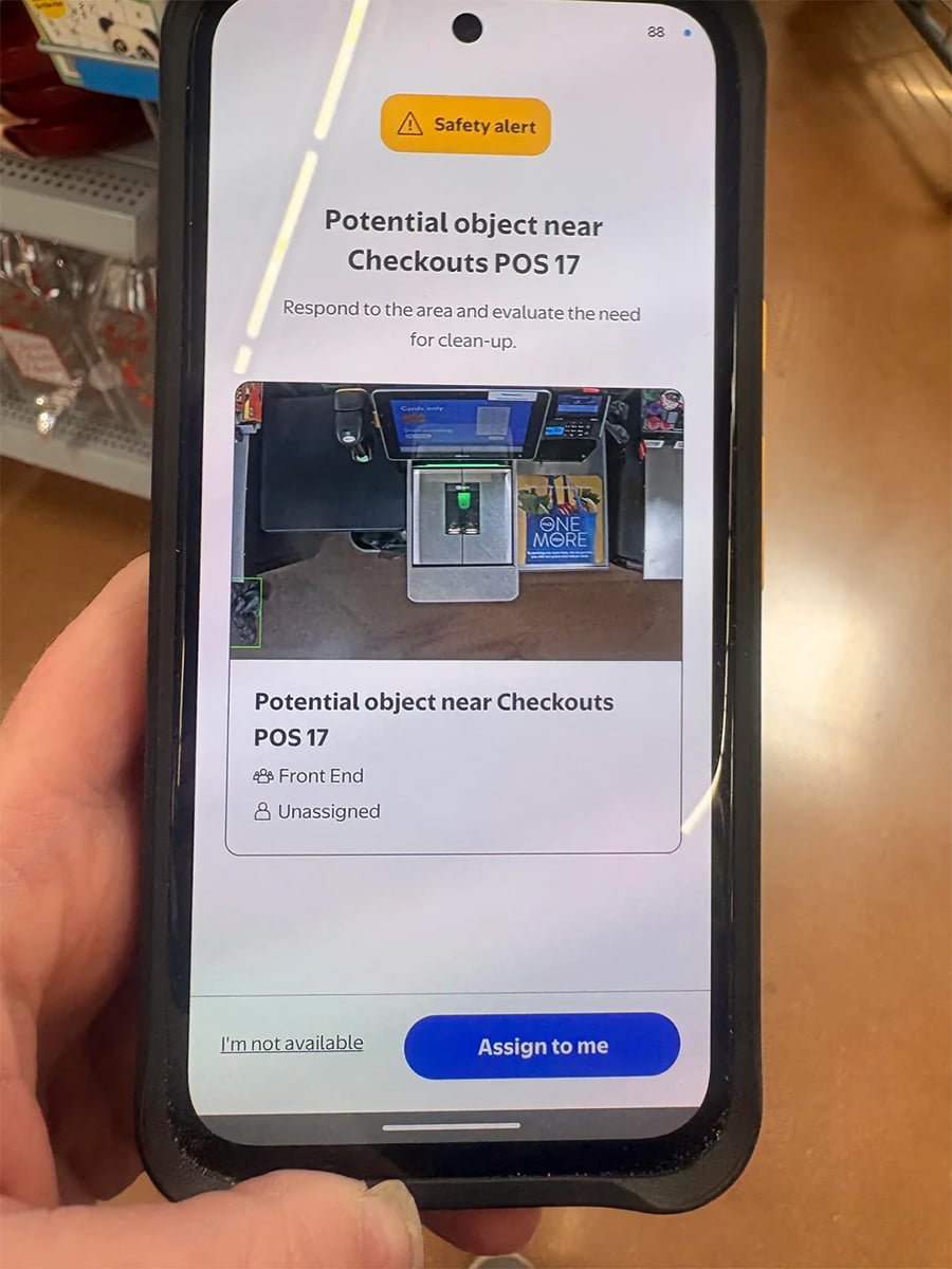 A hand holds a phone displaying a work alert about a potential object near Checkouts POS 17, instructing staff to assess and clean the area.