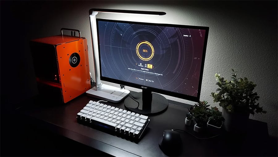 A modern computer setup with a monitor displaying a loading screen, white keyboard, black mouse, orange PC case, desk lamp, and small potted plants on a dark desk.