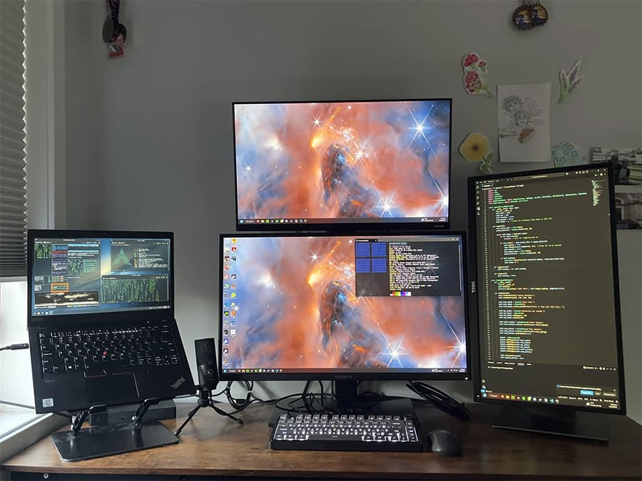A desk setup with three monitors and a laptop. Two monitors display a cosmic wallpaper, one is vertical showing code, and the laptop screen shows graphs. A keyboard, mouse, and some decor are also on the wooden desk.