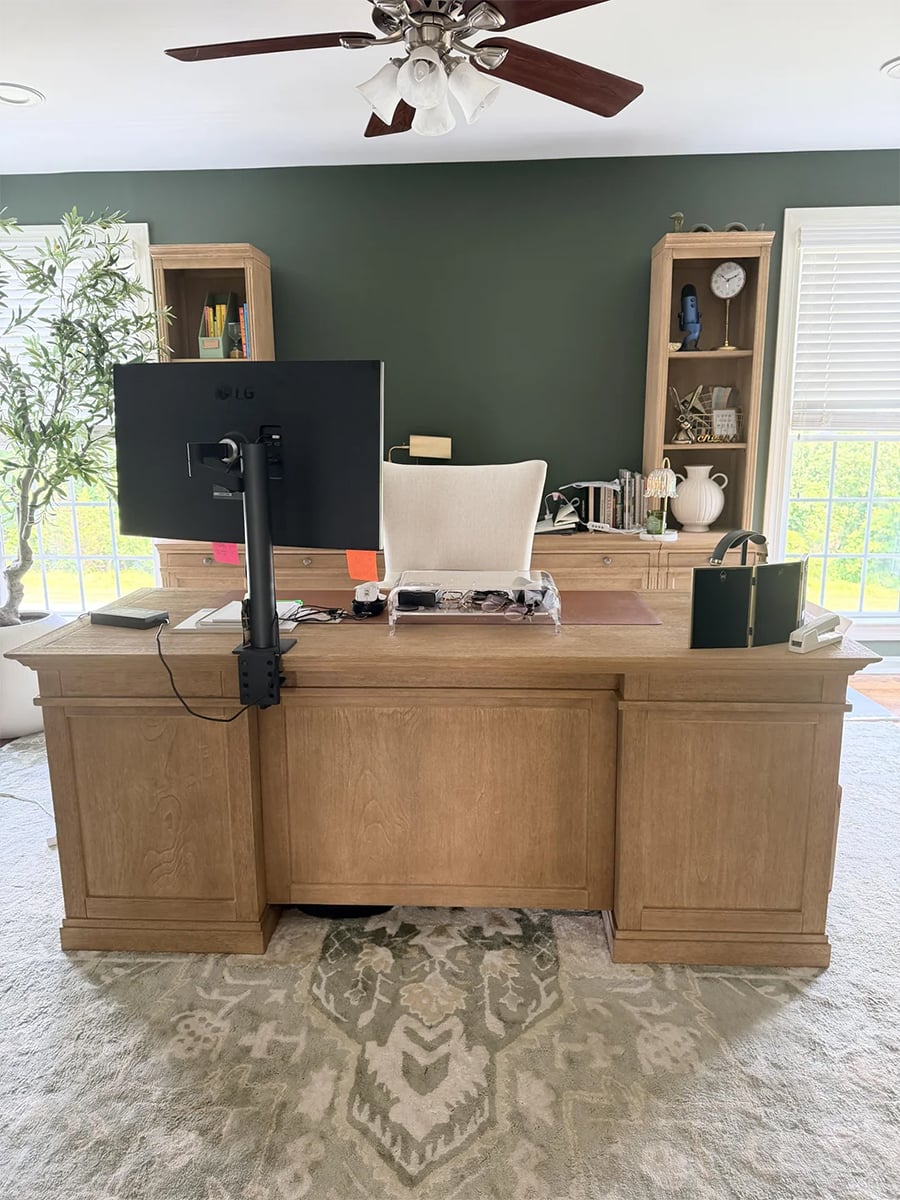 Home office with a large wooden desk, computer monitor, white chair, bookshelves, potted plant, and a ceiling fan. Sunlight streams through windows with white blinds on the green accent wall.