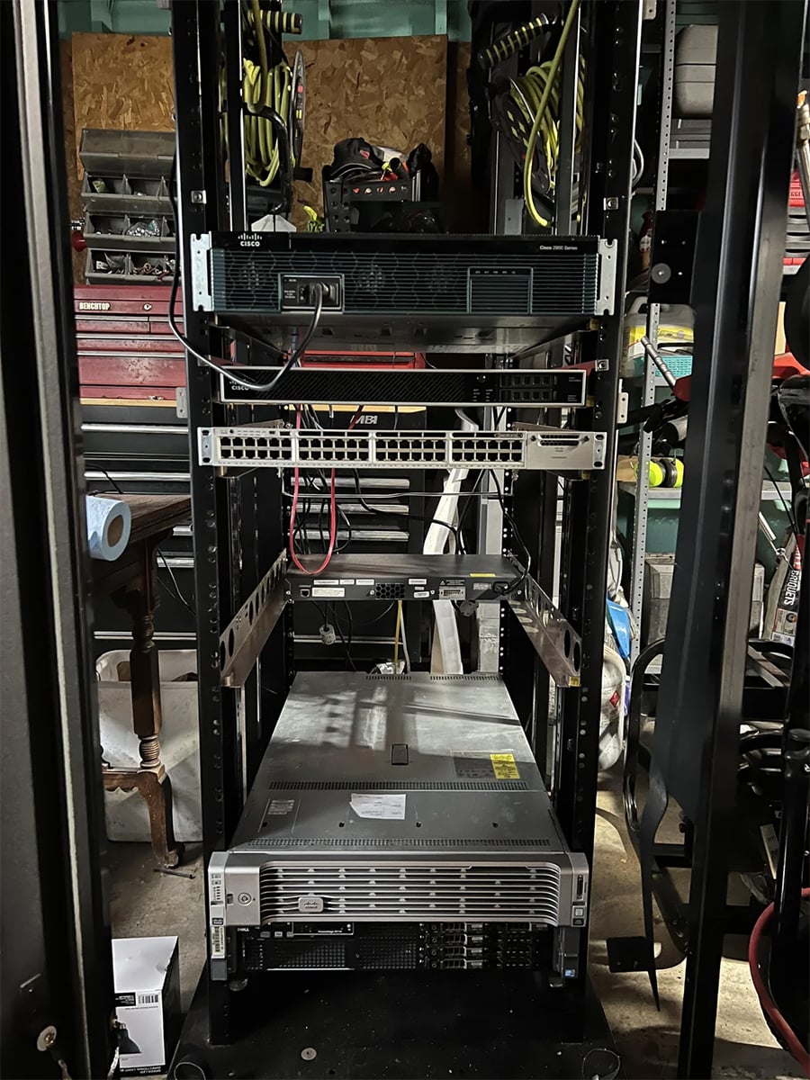 A server rack with several network and server devices, cables, and power cords is set up in a cluttered workshop or garage with tools and shelves visible in the background.