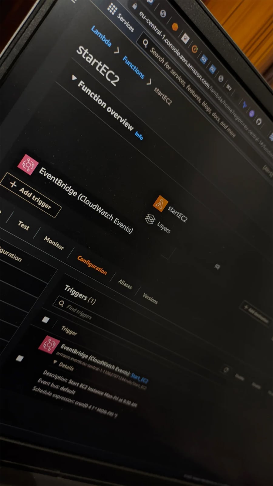A laptop screen displays the AWS Lambda console, showing the configuration page for a function called “startEC2” with EventBridge (CloudWatch Events) set as a trigger.