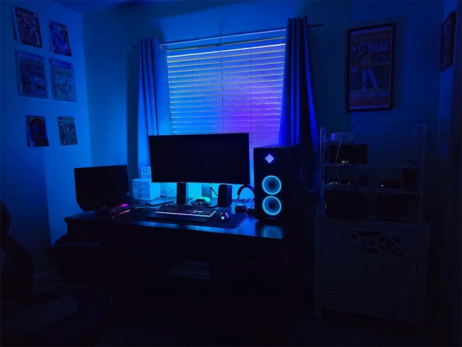 A dark room with a desk setup featuring two monitors and a PC with glowing blue lights, illuminated by blue and purple LED lighting near a window with closed blinds.