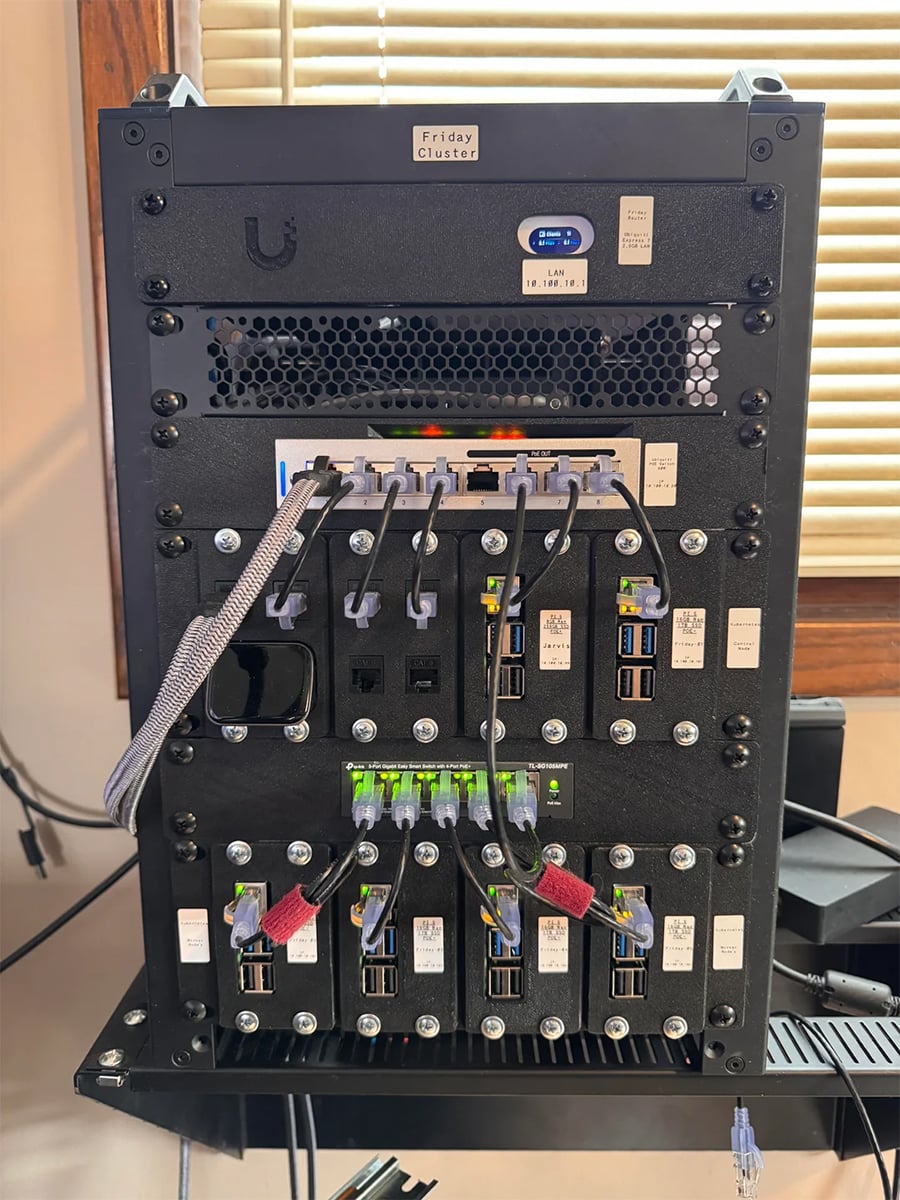 A black computer server rack labeled “Friday Cluster,” with network cables, ports, and indicator lights, set near a window with blinds in the background.