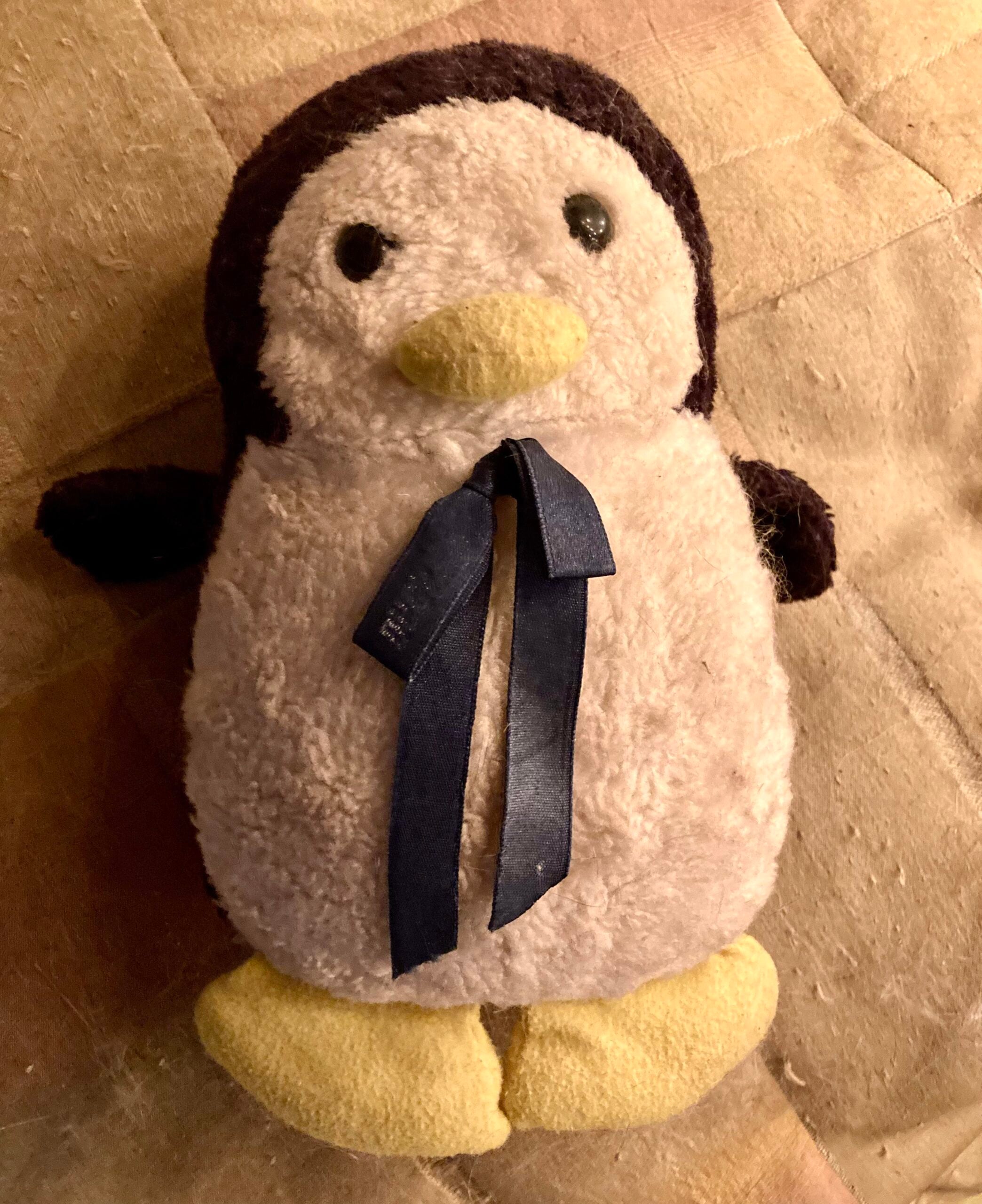 A plush penguin toy with a blue ribbon around its neck lies on a tan, quilted surface. The penguin has yellow feet, a yellow beak, and dark brown wings and head.