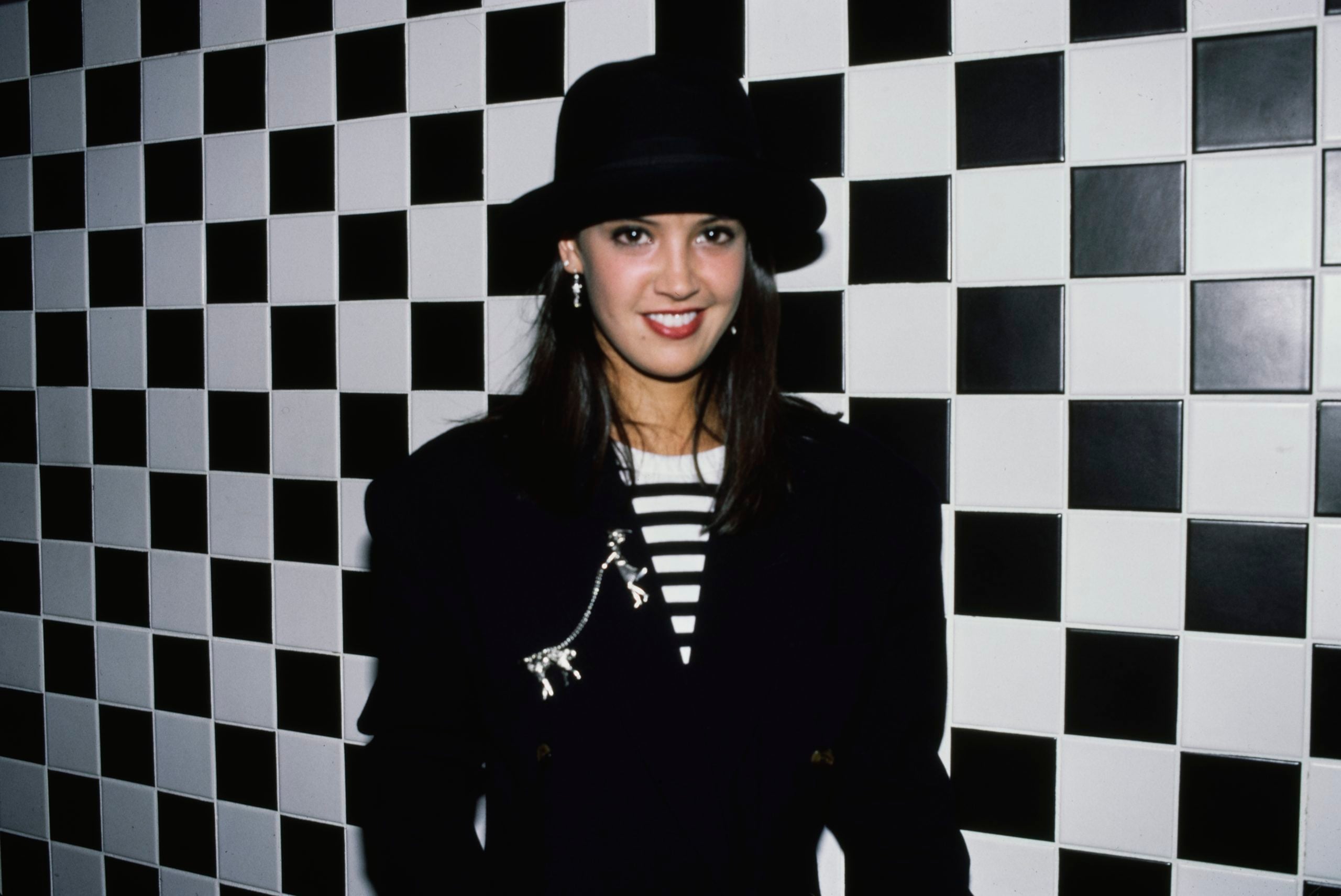 A woman wearing a black hat and dark blazer with a striped shirt stands in front of a black-and-white checkerboard wall, smiling at the camera.