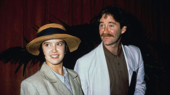 A woman in a straw hat and beige suit stands next to a man with a mustache wearing a light-colored blazer and brown shirt, both looking slightly off camera. A dark background with foliage is behind them.