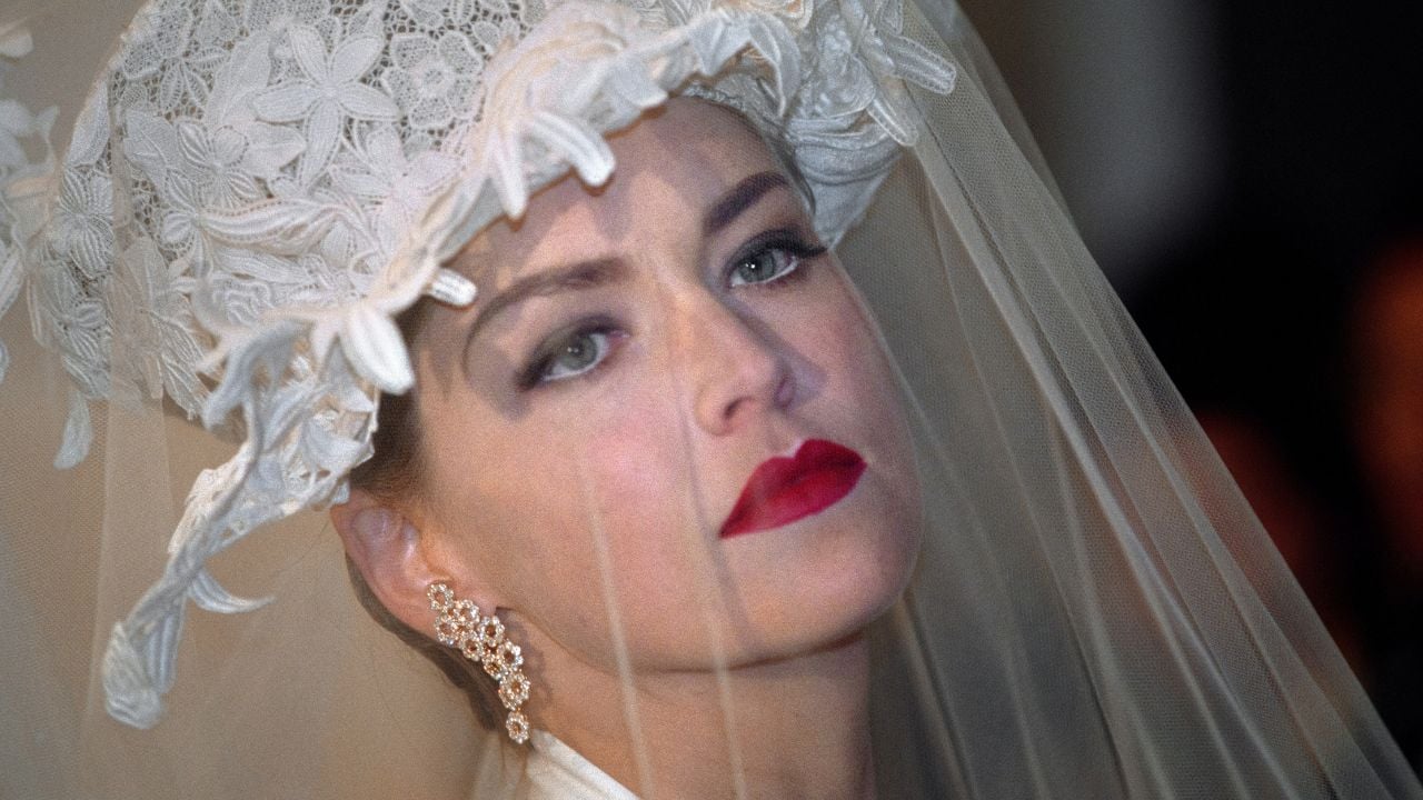 A woman wearing a lace wedding veil and ornate white headpiece with floral details, red lipstick, dramatic eye makeup, and sparkling earrings gazes upward solemnly.