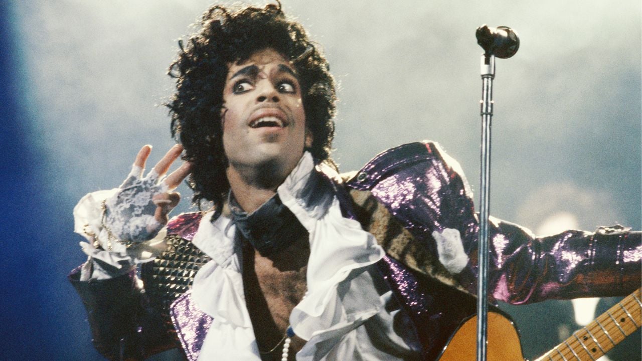 A musician in a ruffled white shirt and shiny purple jacket performs energetically on stage, holding a guitar and gesturing with one hand near his face in front of a microphone.