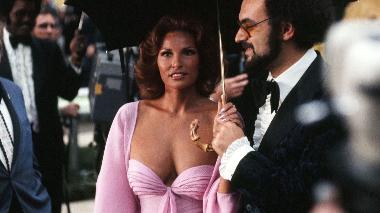A woman in a pink dress with a unique neckline stands beside a man in a suit and glasses who is holding a black umbrella over her at a formal outdoor event.