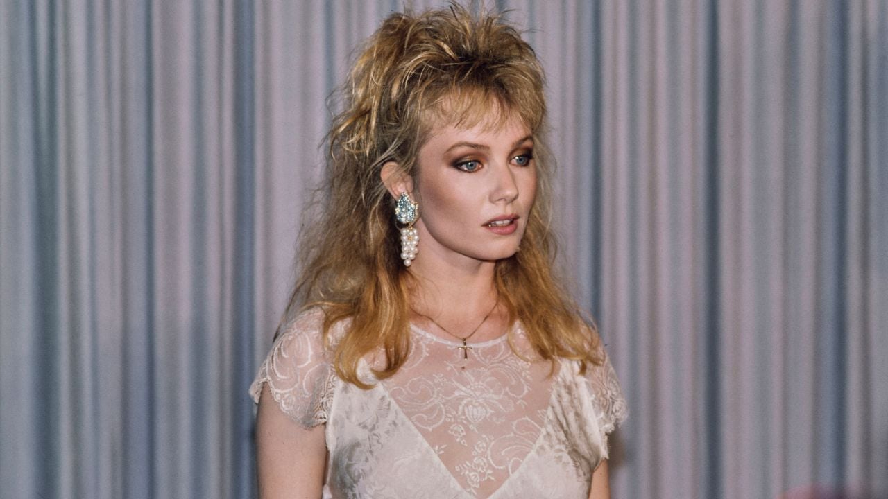 A blonde woman with a voluminous 1980s hairstyle, wearing a white lace dress, large pearl earrings, and a small cross necklace, looking off to the side against a pleated curtain backdrop.