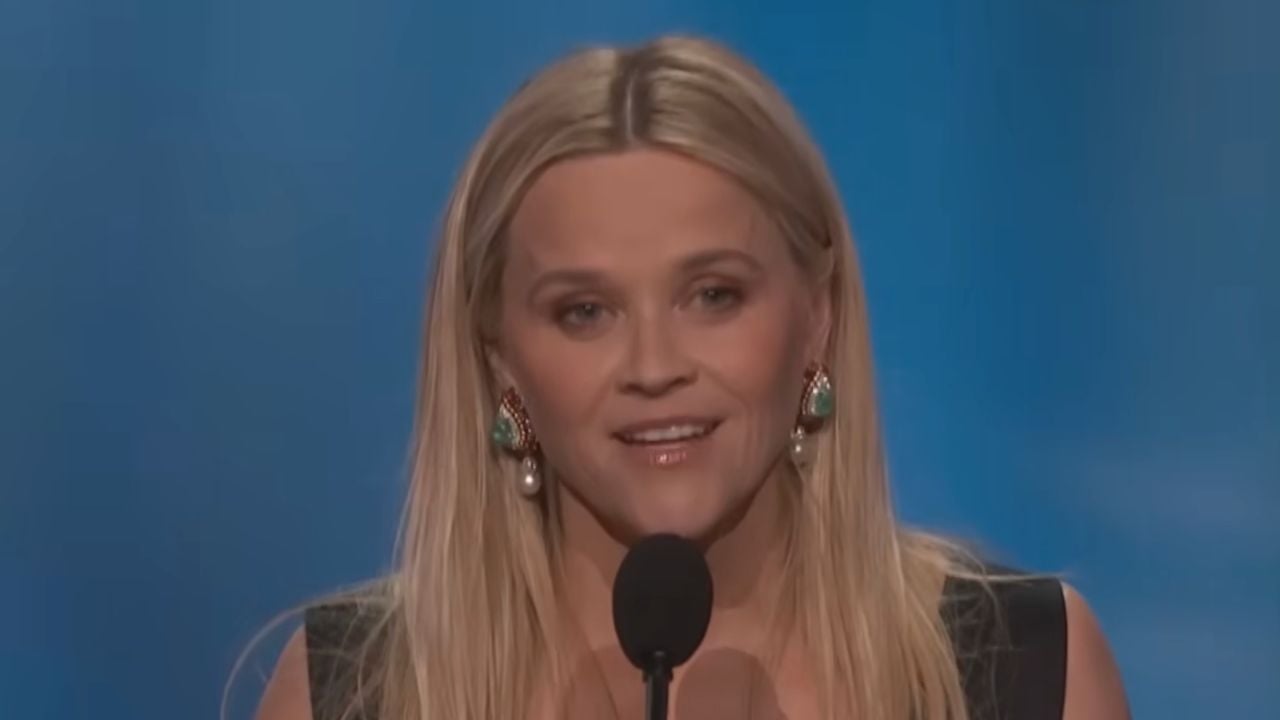 A woman with long blonde hair speaks at a microphone against a blue background, wearing a dark top and statement earrings.