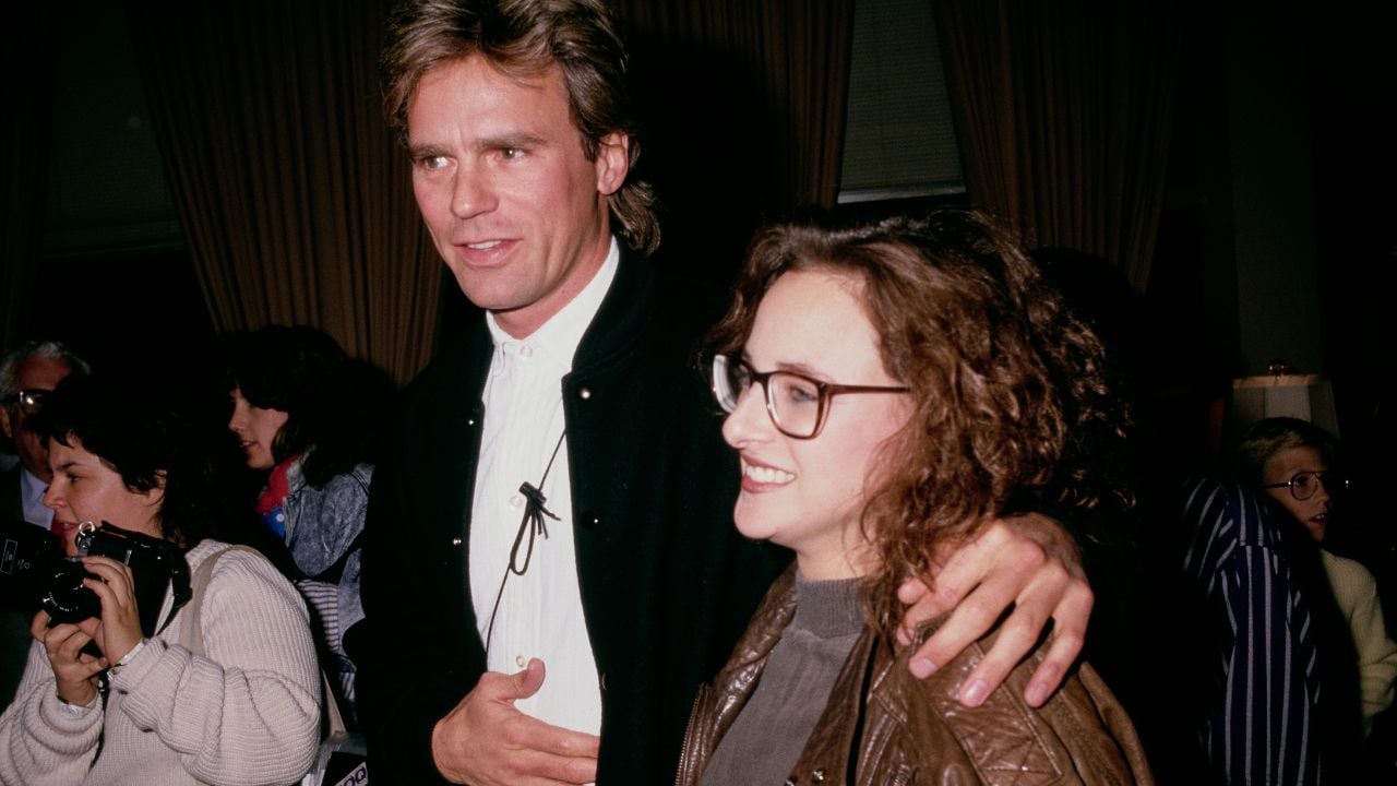 A man with light brown hair and a woman with curly hair and glasses pose together at an indoor event. The man has his arm around the woman. Other people and photographers are visible in the background.